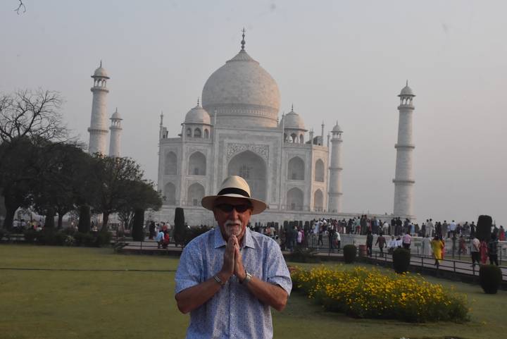 Smiling traveller posing with hands in prayer in front of the hazy white marble Taj Mahal.