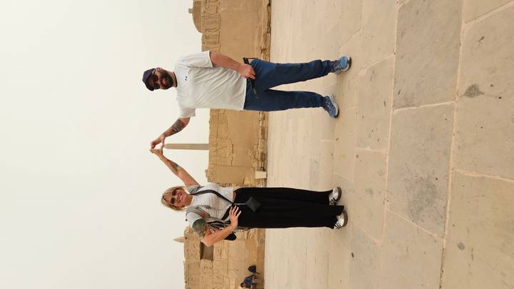 Two travellers perform a playful perspective pose touching the tip of a distant stone obelisk in an ancient temple courtyard.