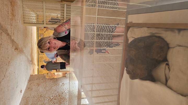 Inside a dimly lit tomb, a smiling visitor looks through a glass case displaying ancient artifacts and painted walls.