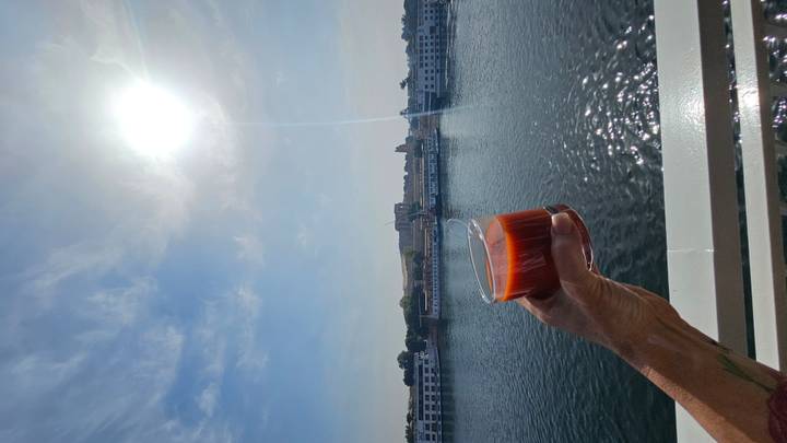 A hand raises a cocktail glass against bright sunlit skies over a calm river lined with cruise ships.
