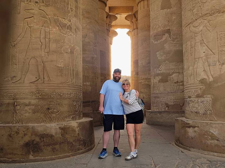 A couple embraces between massive stone columns etched with ancient hieroglyphs, light glowing from an opening behind.