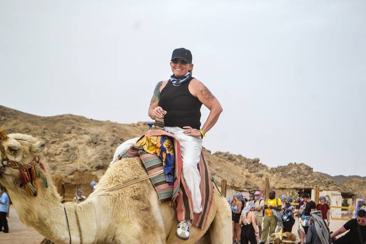 A traveller smiles while riding a camel outfitted with colorful blankets in a rocky desert camp.