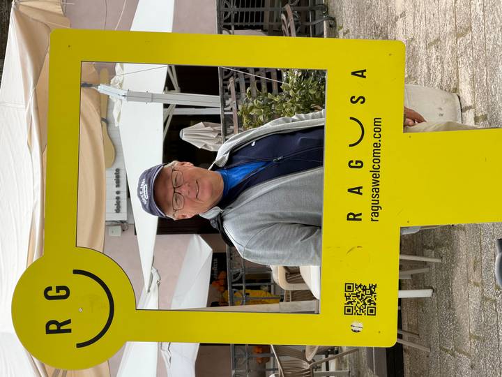 A man smiles from within a bright yellow photo frame advertising Ragusa against a street backdrop.