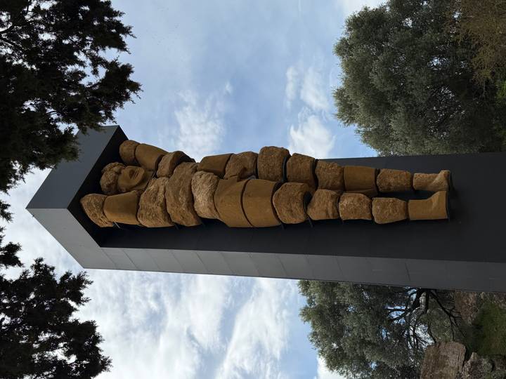 Modern stacked-stone sculpture of an atlas figure stands against a partly cloudy sky amid olive trees.