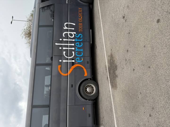 Grey tour bus with large orange and blue 'Sicilian Secrets' branding parked on asphalt.