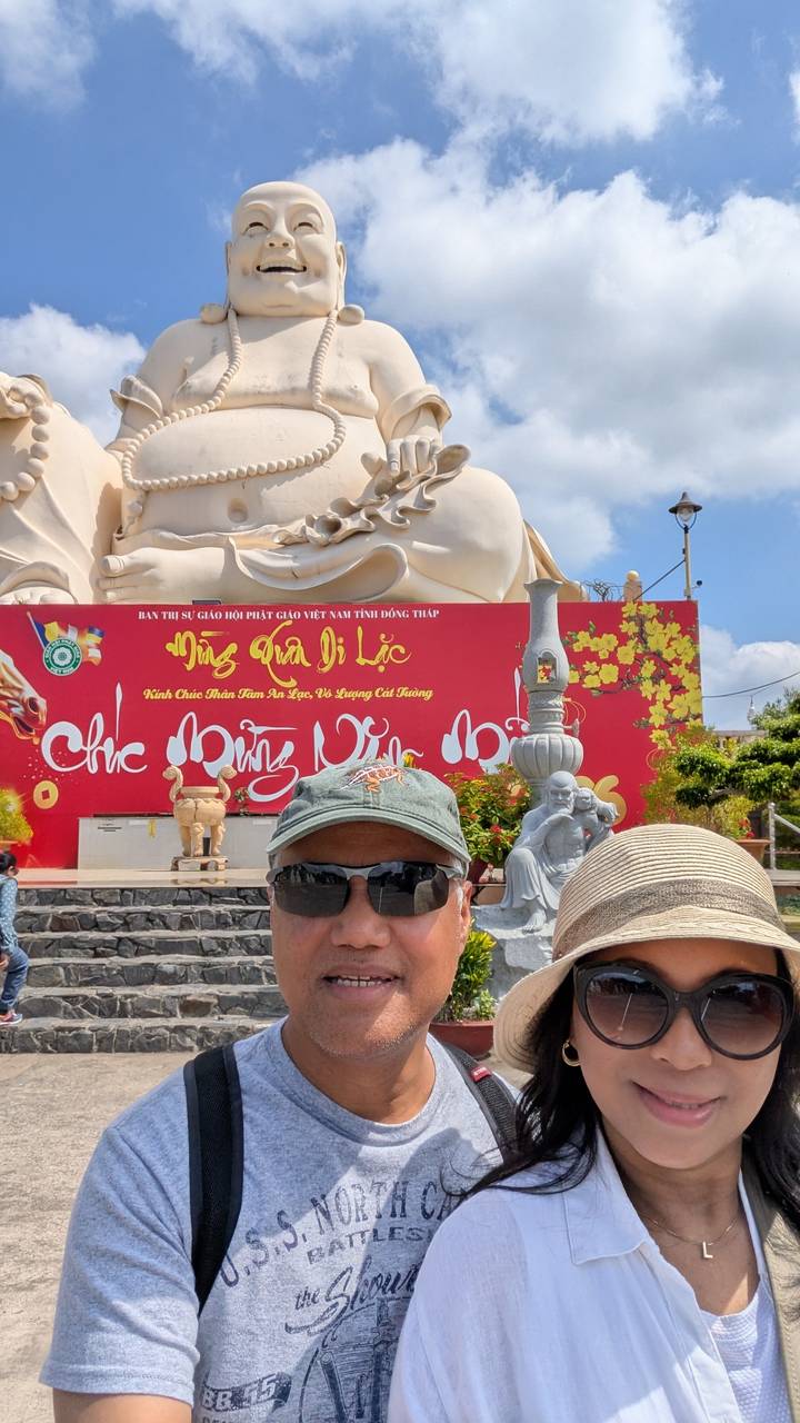 Couple taking a selfie in front of a large white Buddha statue and red festival banner.