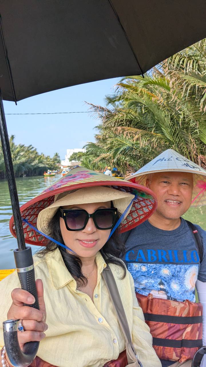 Selfie of two travellers in conical hats on a narrow river surrounded by palms.