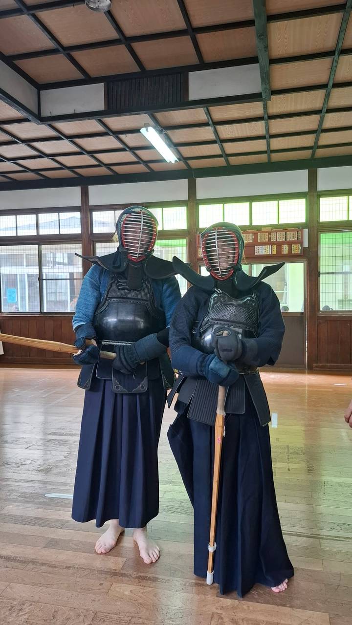 Two people dressed in full kendo armor holding bamboo swords inside a wooden-floored dojo.