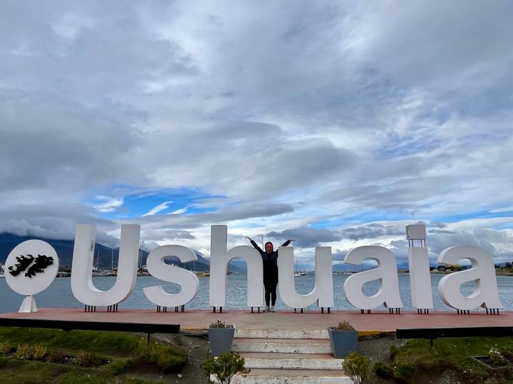 A traveller poses with outstretched arms in front of the oversized white "Ushuaia" sign by the waterfront with mountains and clouds behind.
