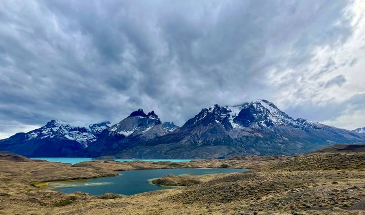 Dramatic snow-capped mountains tower over turquoise lakes and rolling steppe under moody clouds in Patagonia.