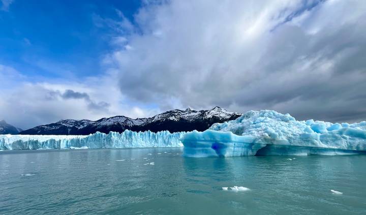 A vast wall of jagged blue ice rises above a calm glacial lake with snowy mountains in the distance.