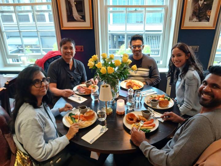 Five travellers share a meal with roast dinners and flowers at a cosy restaurant table.