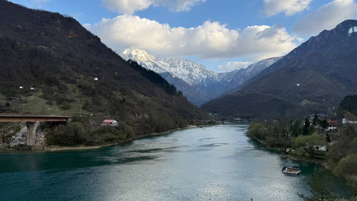 Serene turquoise river winding through a steep mountain valley with snowy peaks under scattered clouds.