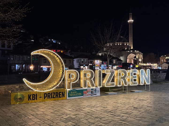 Illuminated crescent-moon and PRIZREN sign shining in a central square at night with a mosque in the background.