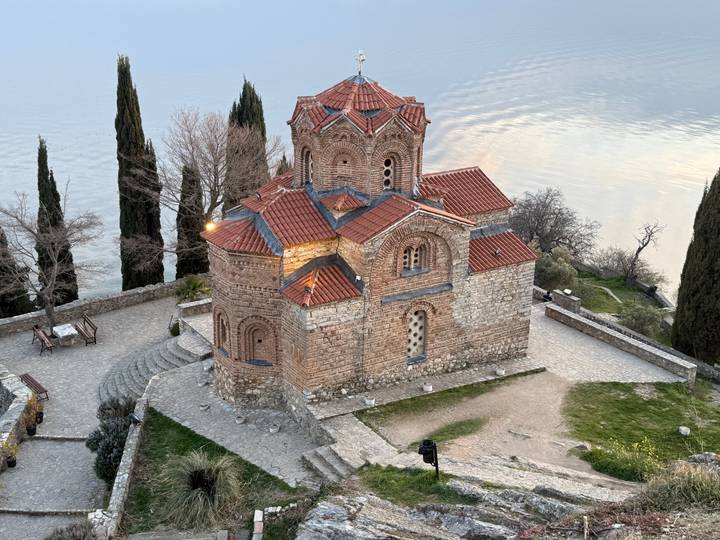 Stone Byzantine church overlooking a calm lake with tall cypress trees and pastel sunset reflections.