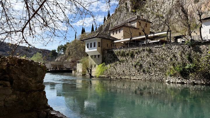 Historic riverside complex of stone buildings and cypress trees mirrored in crystal clear water under a bright sky.