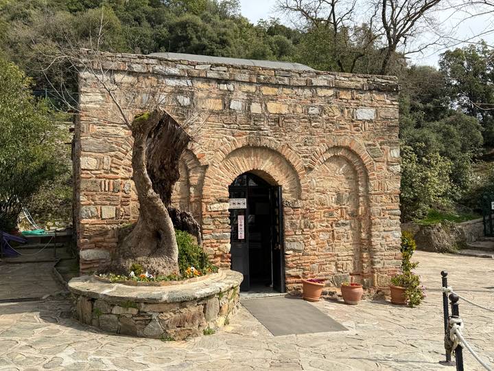 Small stone chapel with twin arched doorway surrounded by potted plants and a gnarled tree trunk.
