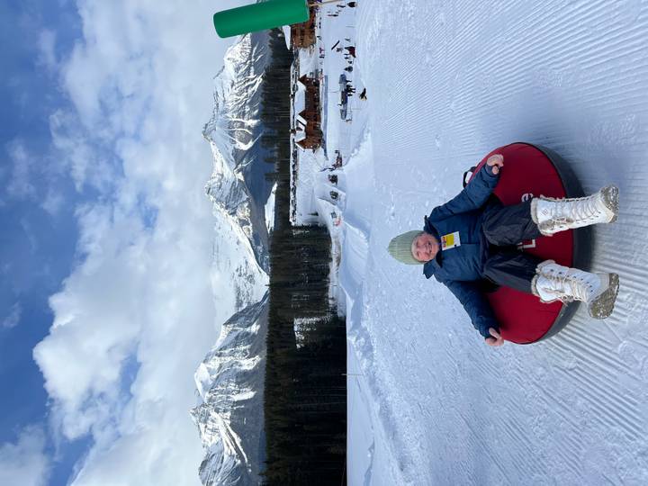 Elderly woman tubing on a groomed snow slope framed by jagged Rocky Mountain peaks.
