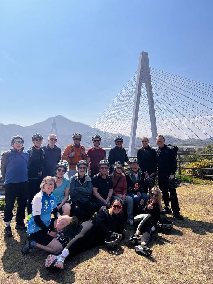 Cycling group poses in front of a cable-stayed bridge with mountain backdrop on a sunny day in Japan.
