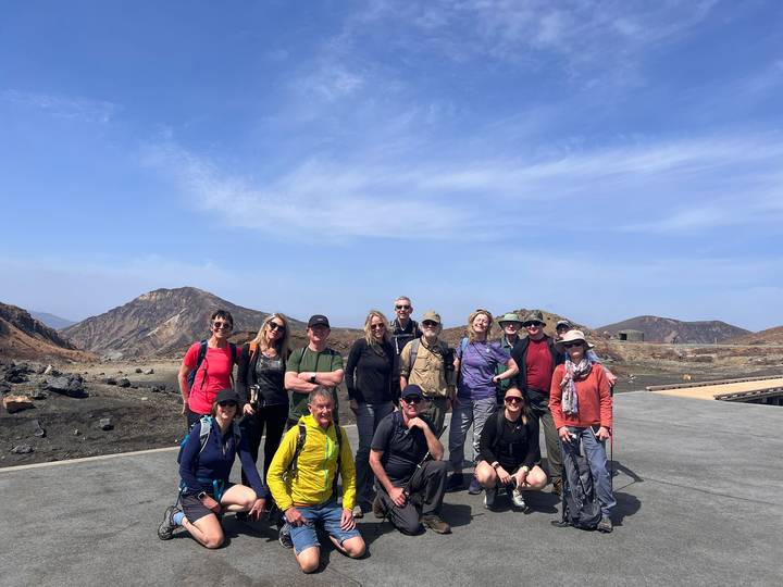 Hiking group poses on a volcanic plateau surrounded by barren peaks under clear blue sky.