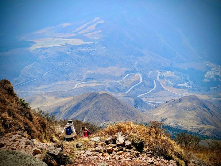 Hikers descend a mountain ridge with winding roads and valleys sprawling far below.