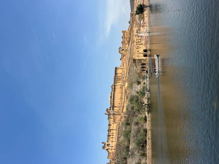 Amber Fort sits majestically above a tranquil lake while a small tourist boat crosses the water under clear blue skies.