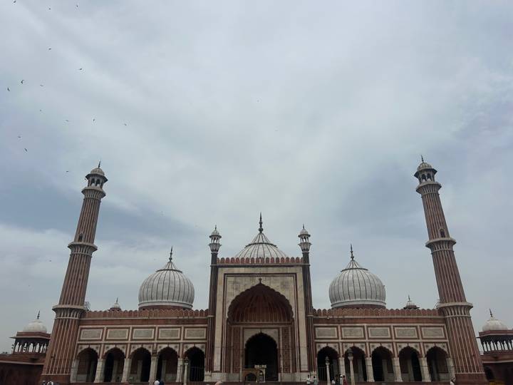 Grand facade of Jama Masjid with three white domes and towering minarets under a cloudy sky.