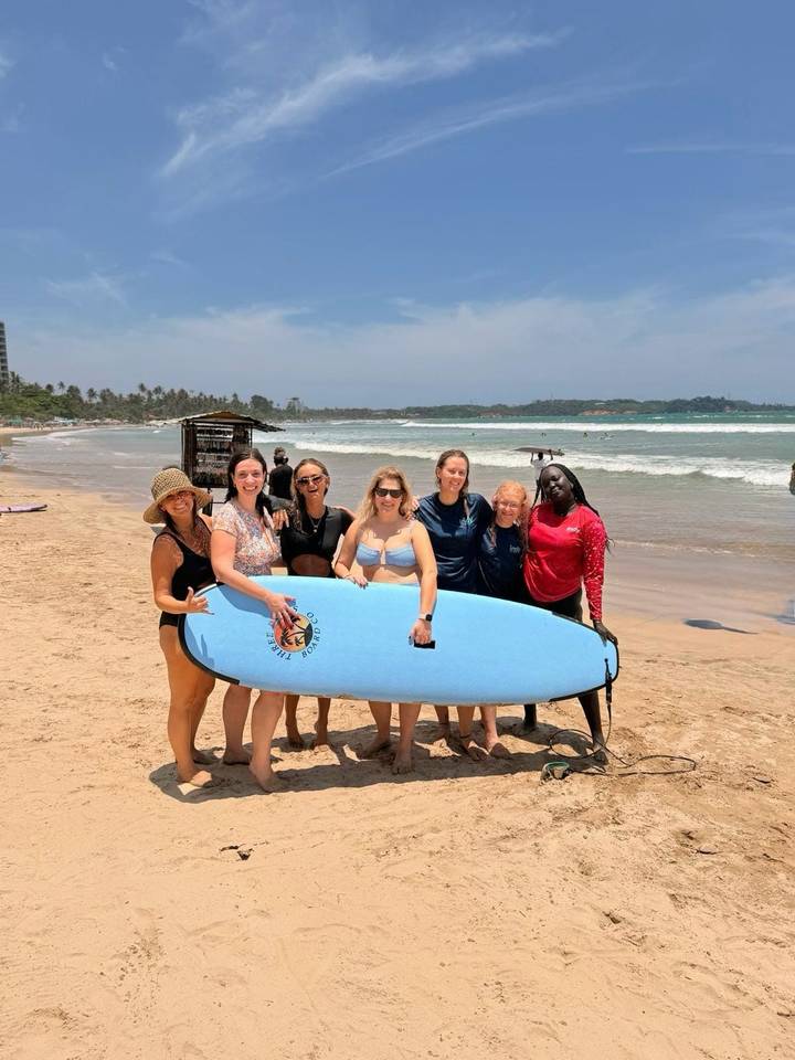 Group of women hold a surfboard and smile on a sunny tropical beach with waves rolling in behind.