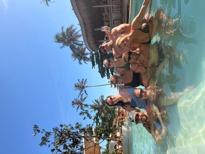 Happy travellers pose together in a sun-lit resort pool surrounded by palm trees.