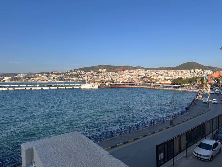 Blue waters of Kusadasi harbour with town buildings spread across the hillside under a clear sky.