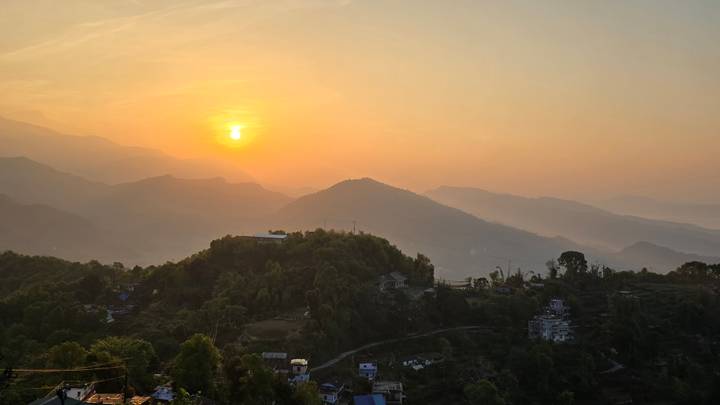 Golden sunrise breaking above layered Himalayan foothills and rural houses.