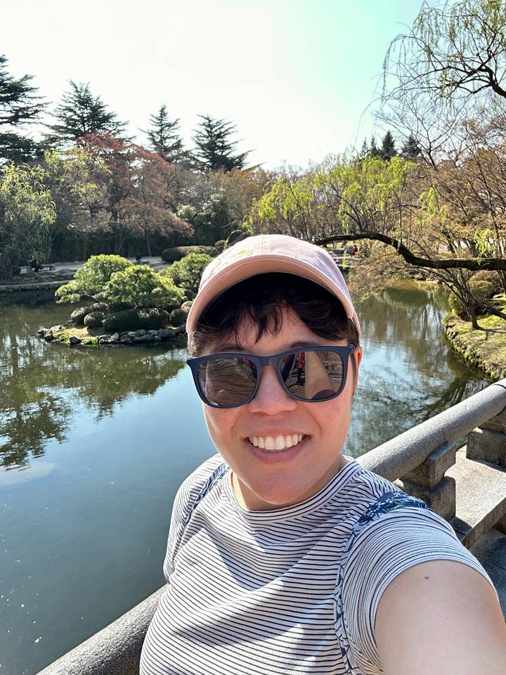 Smiling traveler taking a selfie on a stone bridge over a landscaped pond in a sunny garden.