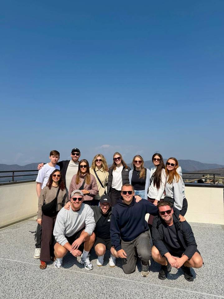 Large group of travelers posing on a terrace with a faint snow-capped Mount Fuji in the background under a clear sky.