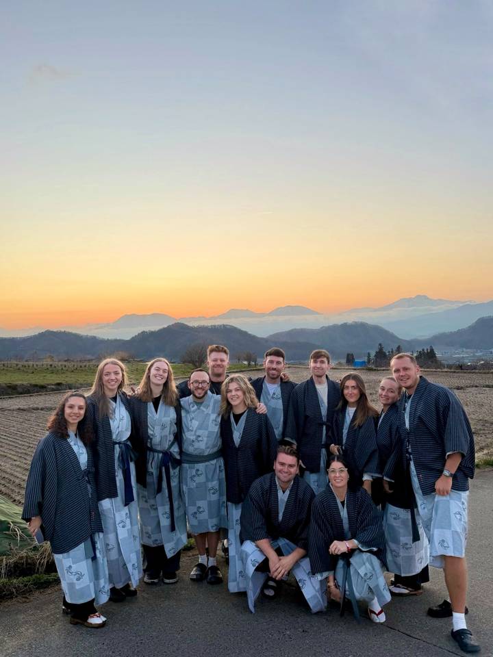 Travel group in matching robes standing in front of layered mountains at sunset with an orange sky.