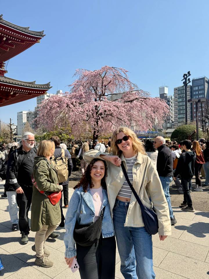Two women smiling under a cherry blossom tree amid crowds at a city temple fair.