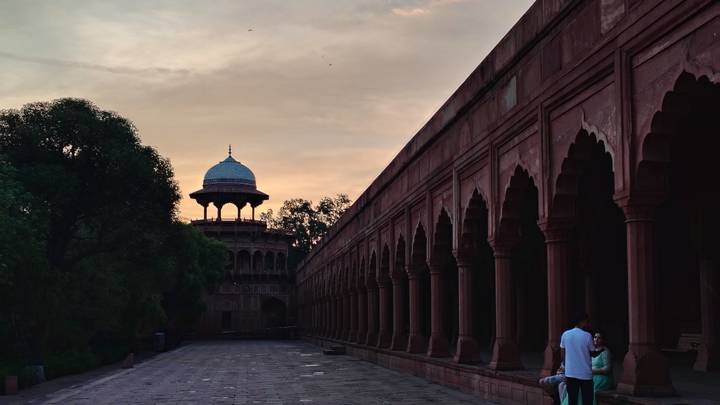 Long colonnade of red sandstone arches at sunset with a domed pavilion silhouetted at the end.