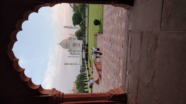 The Taj Mahal framed by a red sandstone archway with visitors walking across the courtyard at dusk.