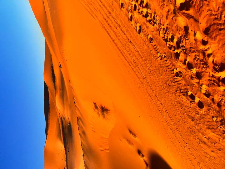 Close-up of rippled desert sand with vehicle tracks and rolling dunes under a bright blue sky.