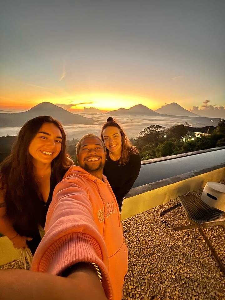 Three smiling travellers take a sunrise selfie above a sea of clouds with volcanic peaks silhouetted in the background.
