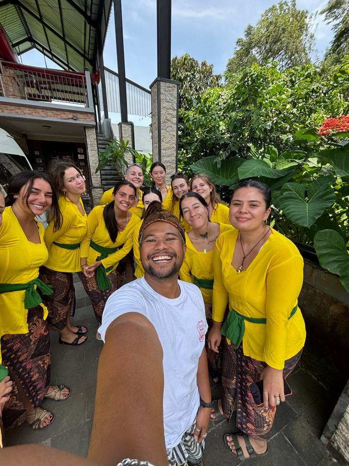A cheerful group dressed in matching yellow traditional outfits takes a selfie in a tropical courtyard.
