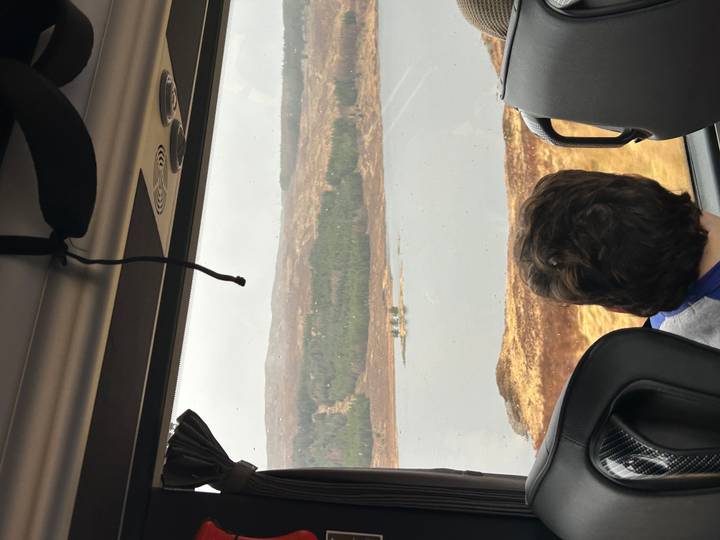 A passenger gazes out a coach window at a remote Scottish loch and forested hills in drizzly weather.