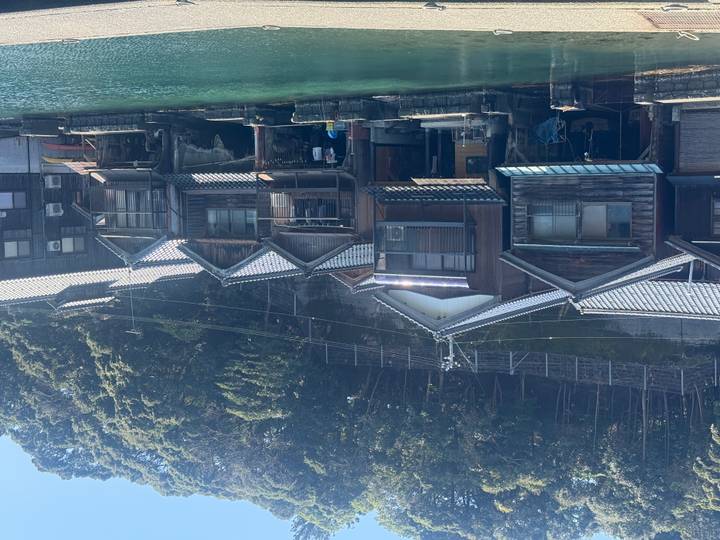 Traditional wooden boathouses line a teal-colored inlet beneath a forested hillside on a bright day.