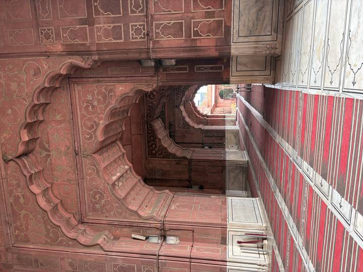 Long arched sandstone corridor with intricate Mughal carvings and striped prayer rugs.