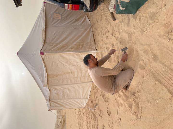 Man kneeling in desert sand hammering a stake to pitch a canvas tent.