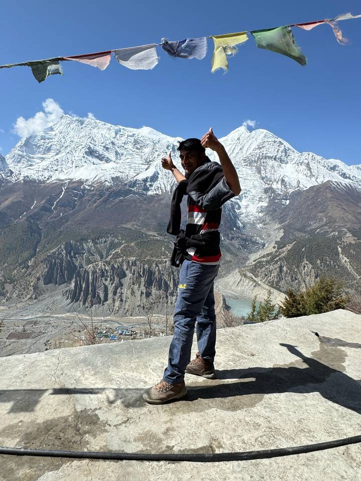 Trekker points triumphantly at the towering snow mountains from a rocky lookout.