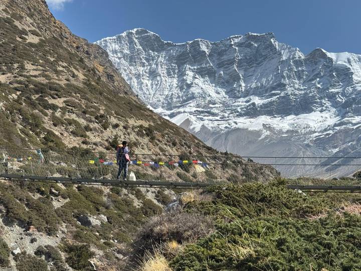 Another view of hiker crossing prayer-flag suspension bridge with glacier-clad peaks beyond.