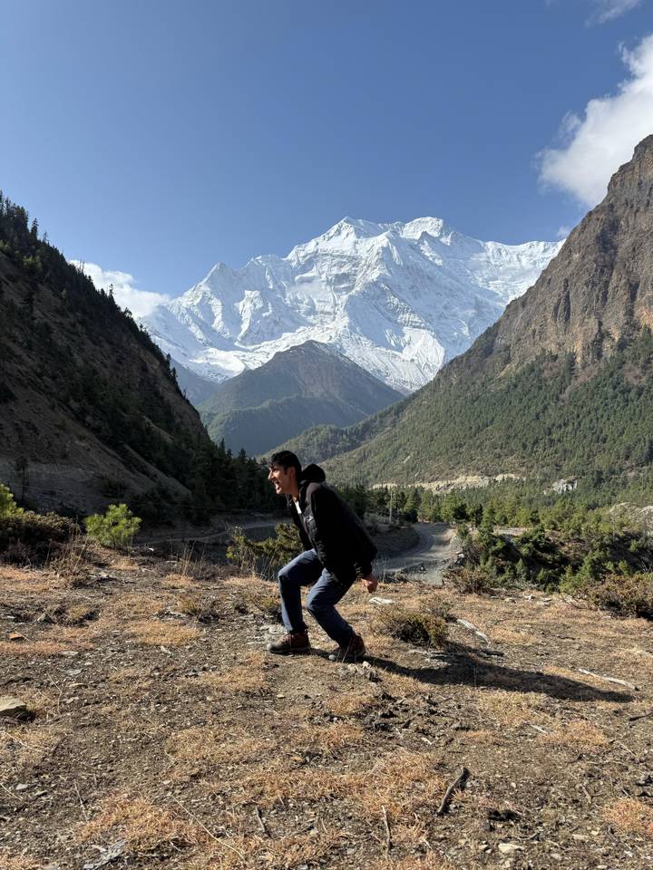 Hiker crouches playfully on mountain viewpoint surrounded by pine forests and snowy peaks.