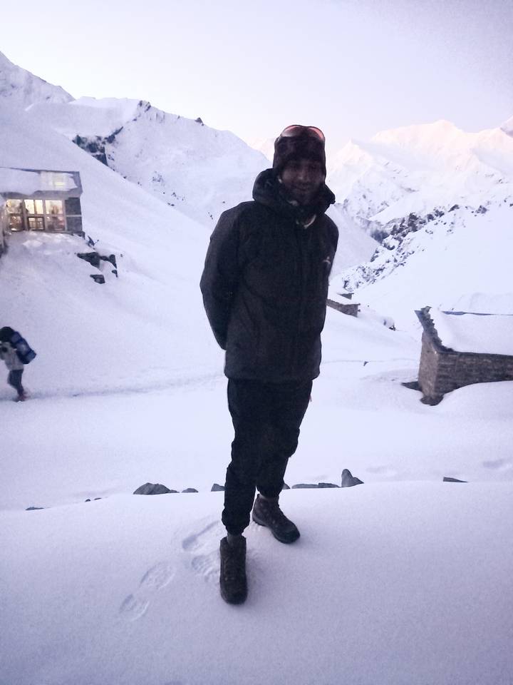 Low-light portrait of trekker standing in deep snow near stone buildings at dusk.
