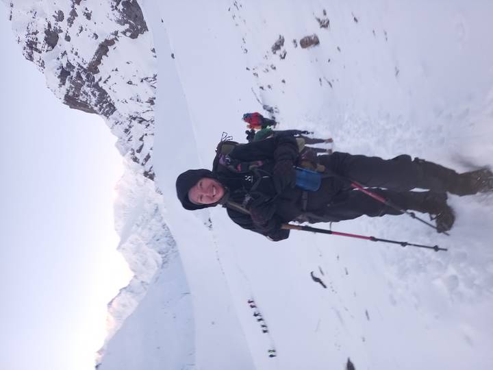 Smiling woman with trekking poles walking up snowy trail toward bright peaks.