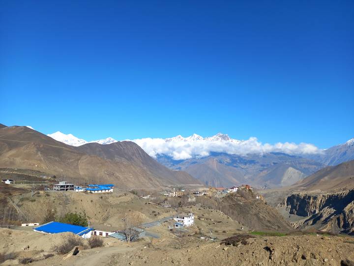 Clear blue-sky panorama of brown valleys and distant snowy Himalayan summits above small villages.